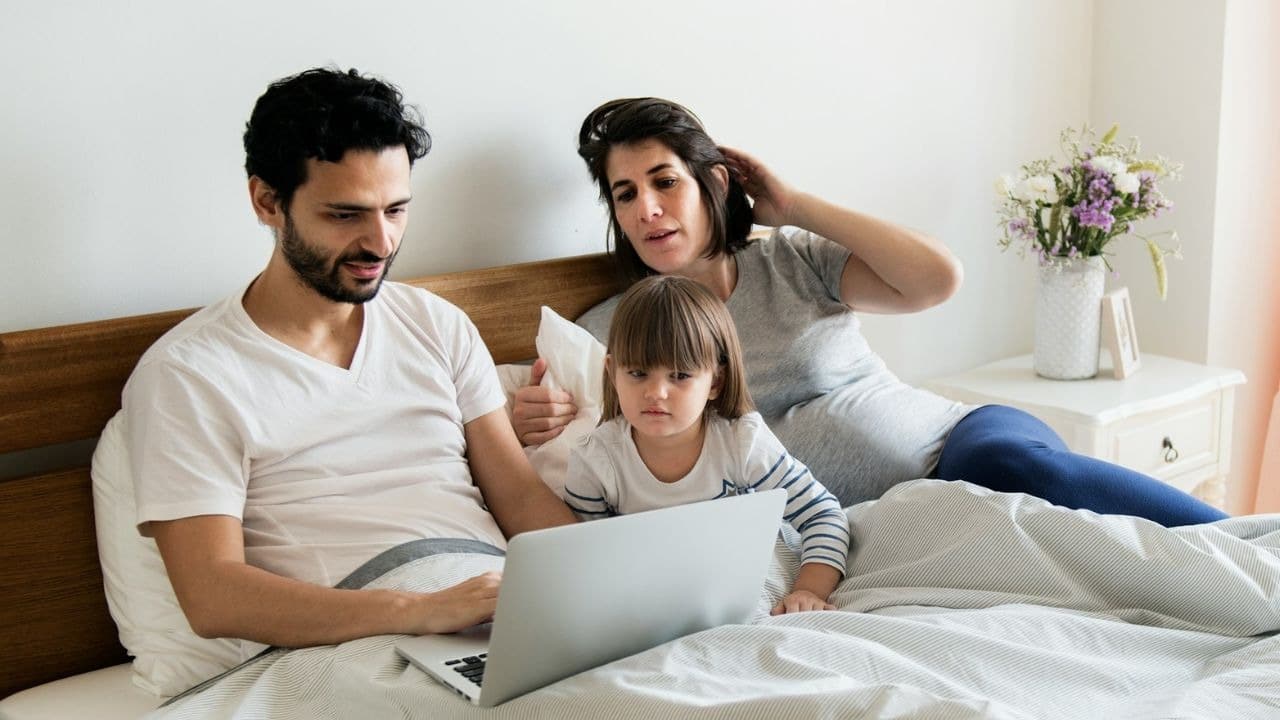 A man, a pregnant woman, and a young child look at a laptop in bed.