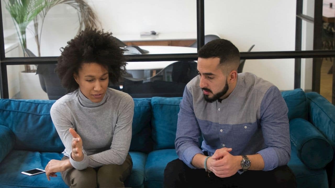 A woman with afro-textured hair and a bearded man sit together on a blue sofa.