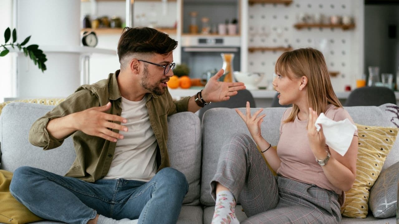 A man and woman sit on a gray sofa, gesturing with their hands while talking.