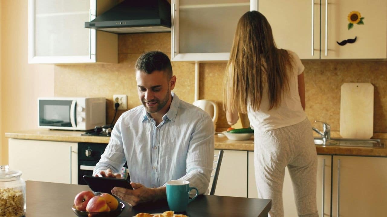 A man uses a tablet at a table while a woman works in the kitchen.
