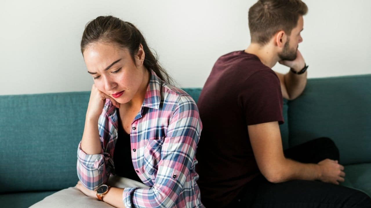 A man and woman sit back-to-back on a blue sofa looking away from each other.