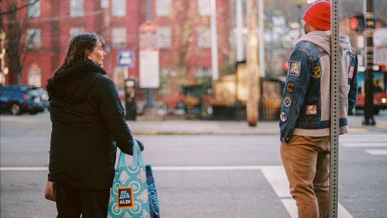 A woman holding a shopping bag stands outdoors looking toward a man in a denim jacket.