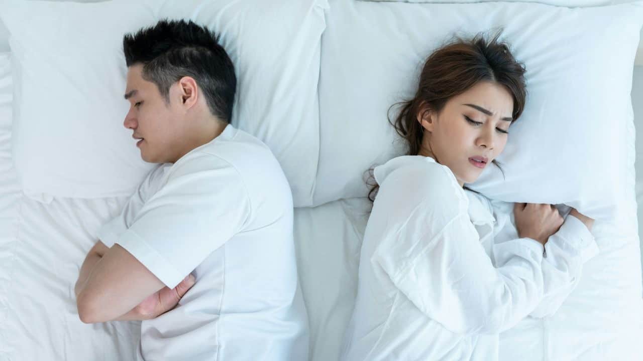 A man and woman lie back-to-back on a white bed with their eyes closed.