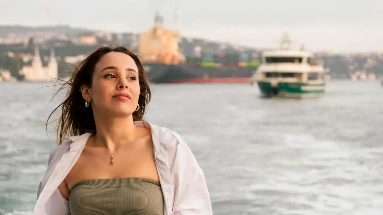 A woman standing by the water with a boat in the background.