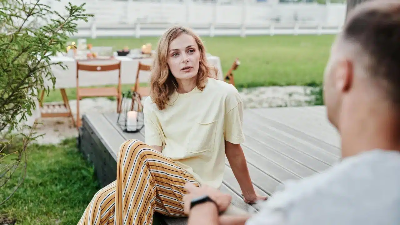 A woman sitting outdoors on a deck, talking to another person.