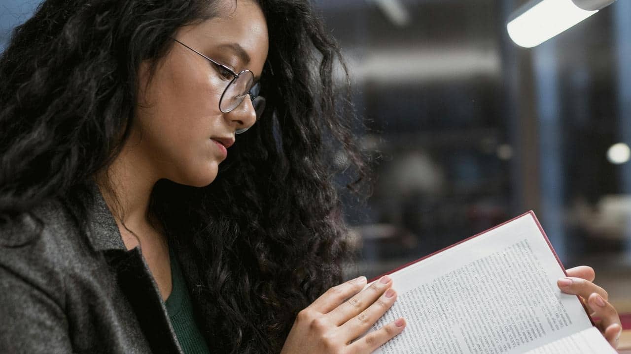 A woman wearing glasses reading a book under a light indoors.