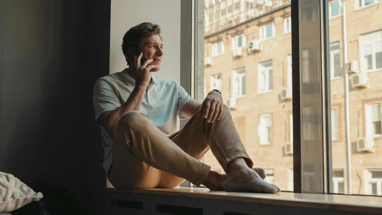 A man sitting by a window talking on a phone.