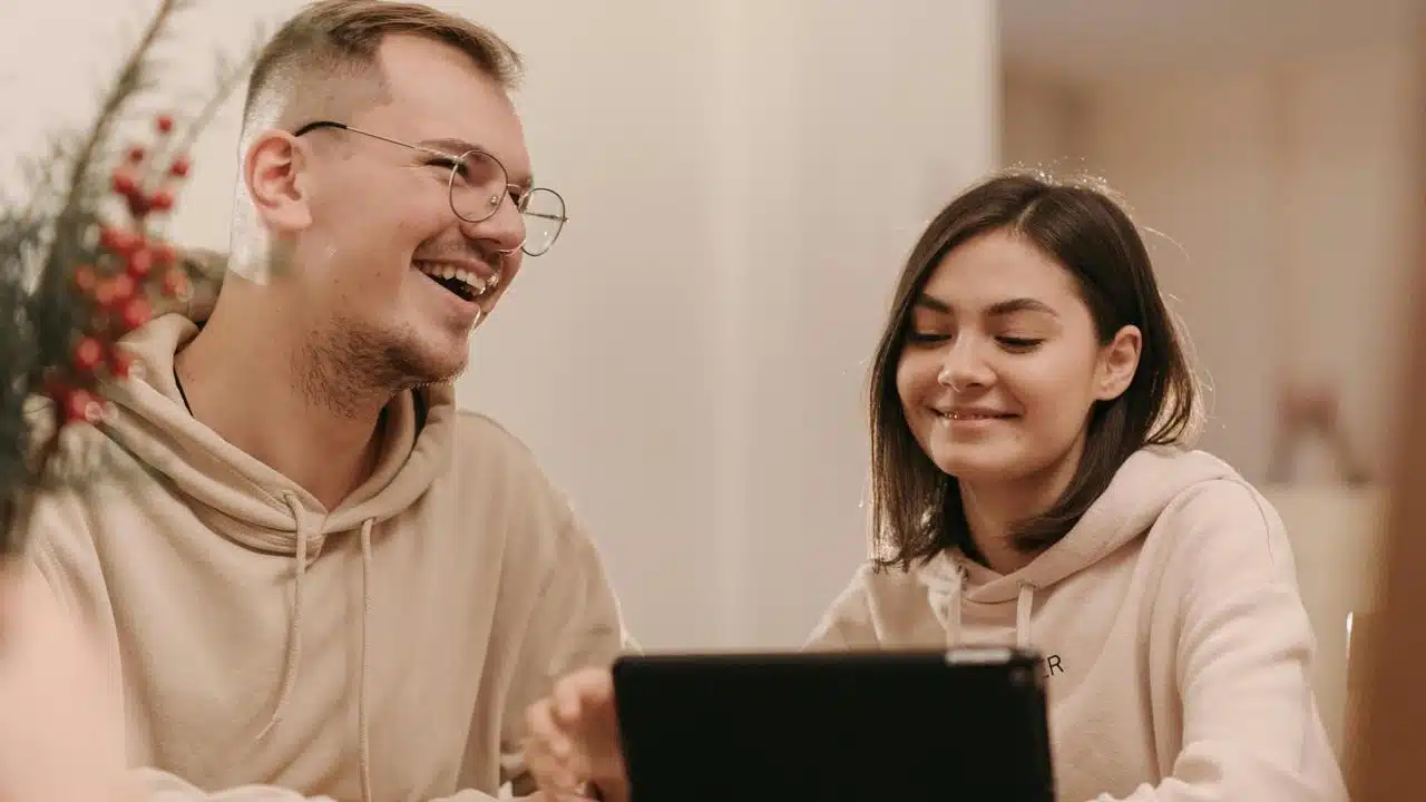 A man and woman smiling while looking at a tablet indoors.