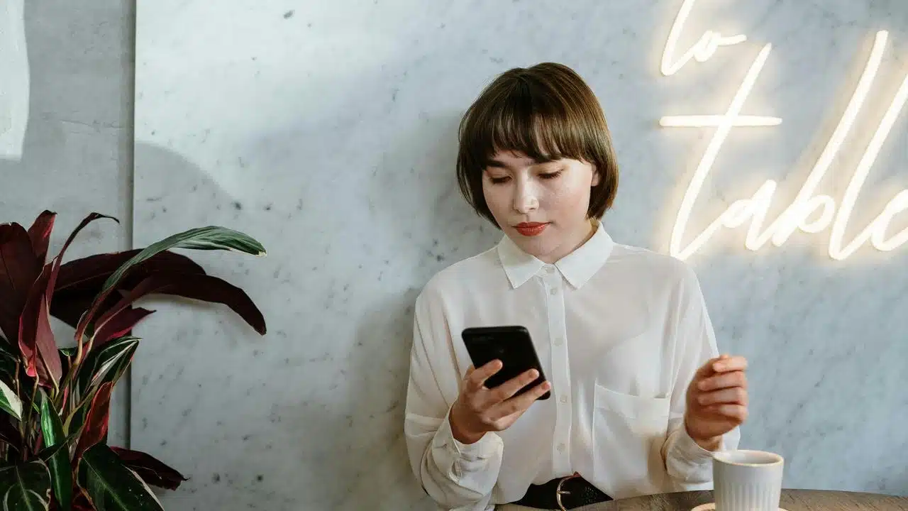 A woman sitting at a table looking at her phone with a cup of coffee nearby.