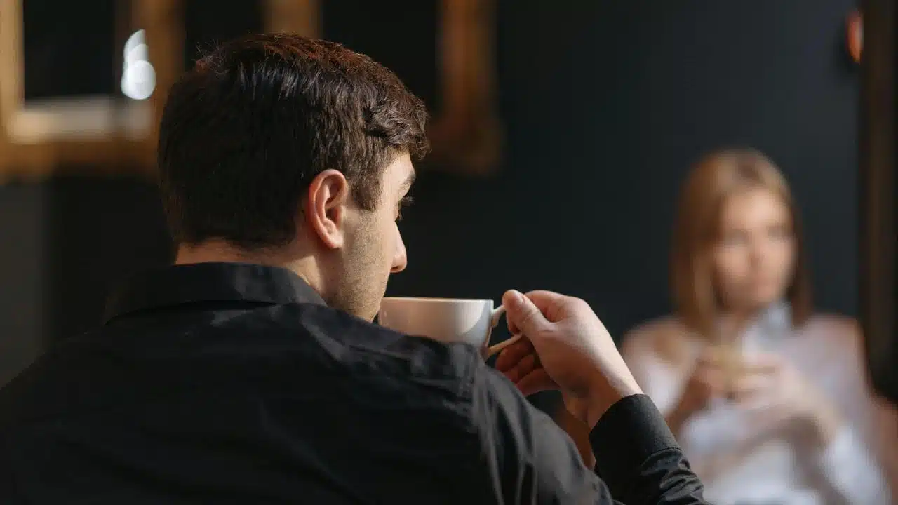 A man drinking coffee while sitting across from a woman in a dimly lit setting.