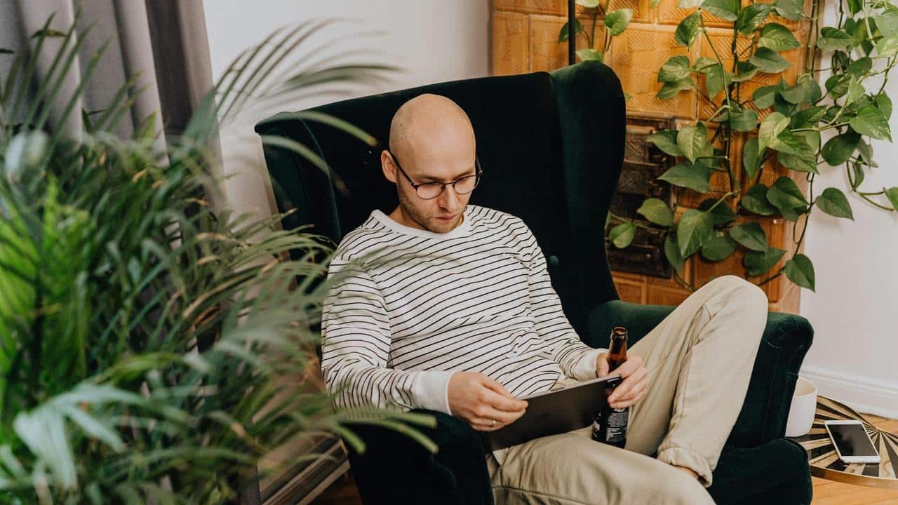 A bald man with glasses sitting in a chair using a tablet while holding a drink.