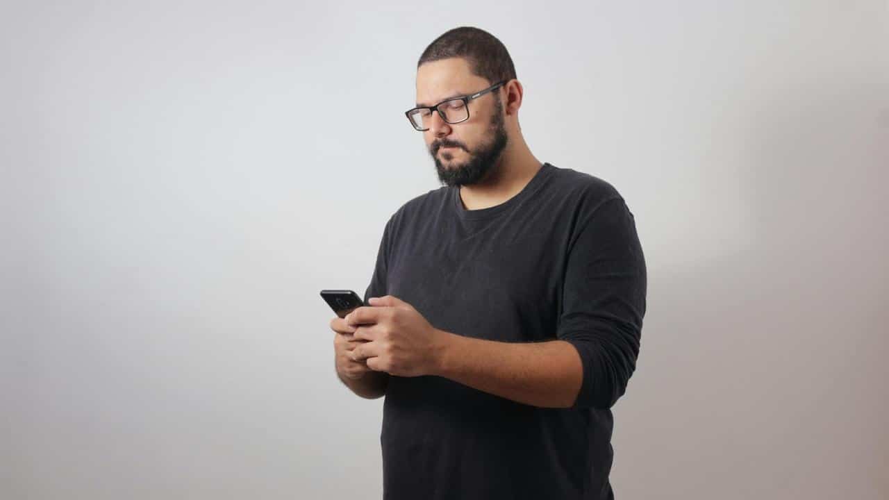 A man wearing glasses looking down at his smartphone against a plain background.