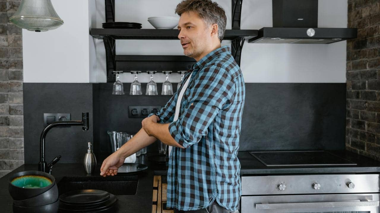 A man in a kitchen rolling up his sleeve near a sink and stove.