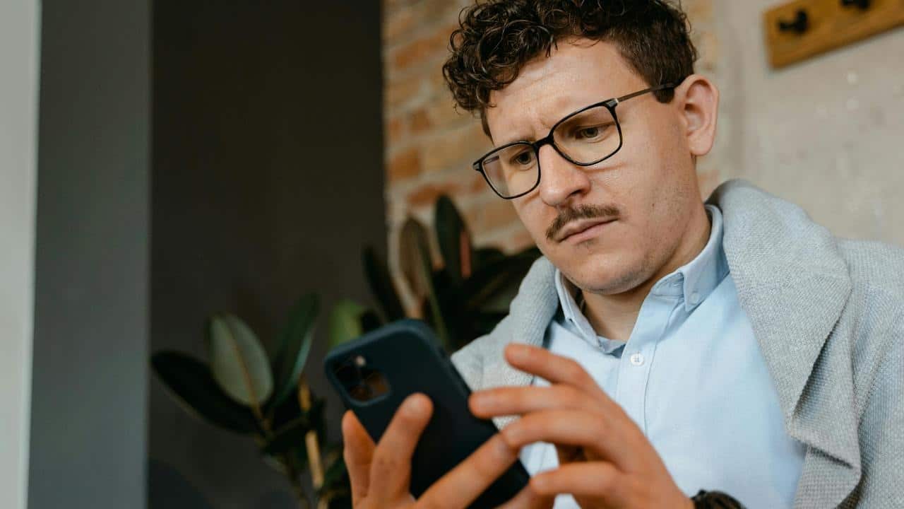 A man with glasses and a mustache looking intently at his smartphone indoors.