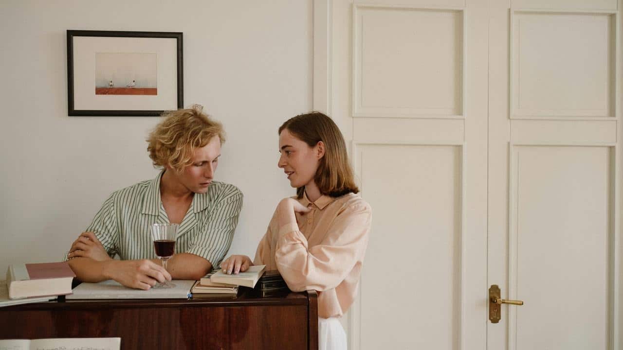 A man and woman leaning on a piano, talking with books and a glass of wine nearby.