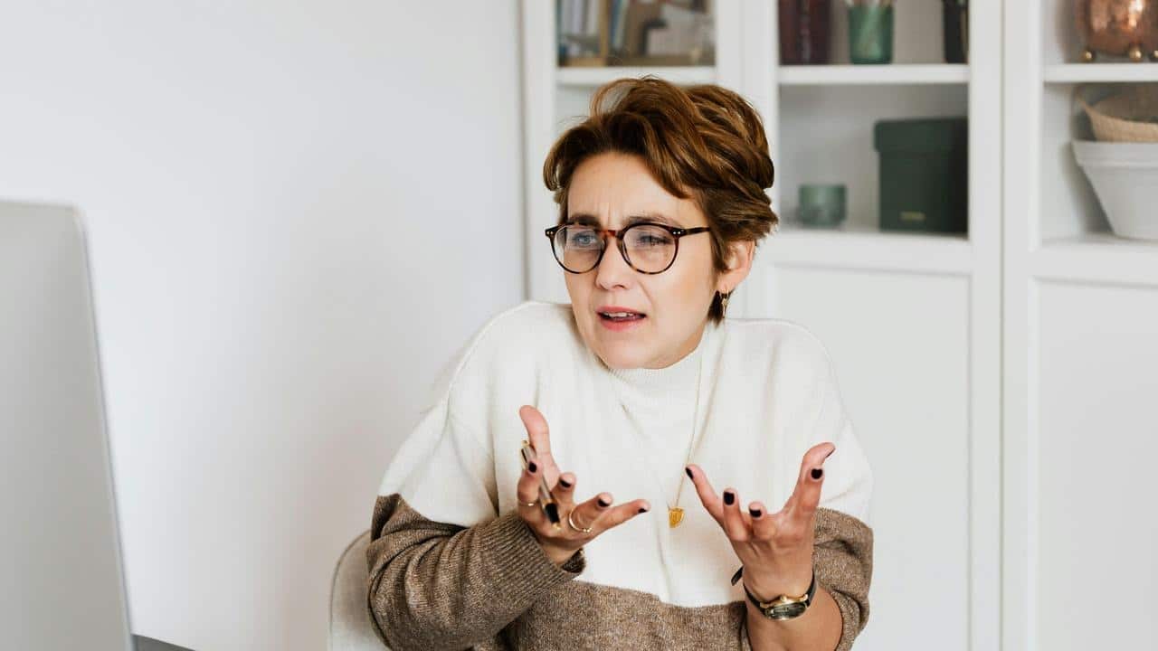 A woman with glasses gesturing in confusion while sitting at a desk.
