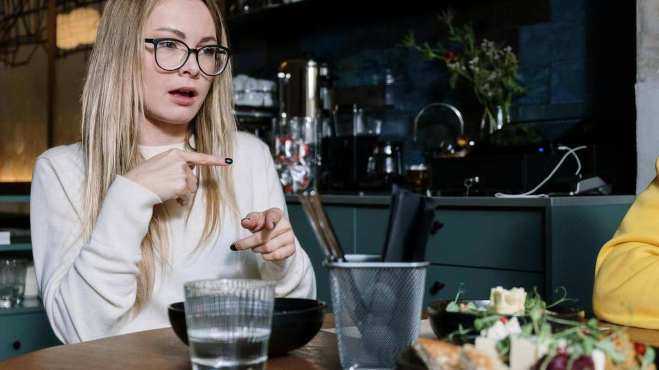A woman with glasses pointing while talking at a dining table.