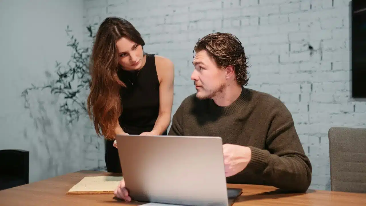 A woman leans over to talk to a man sitting at a desk with a laptop.