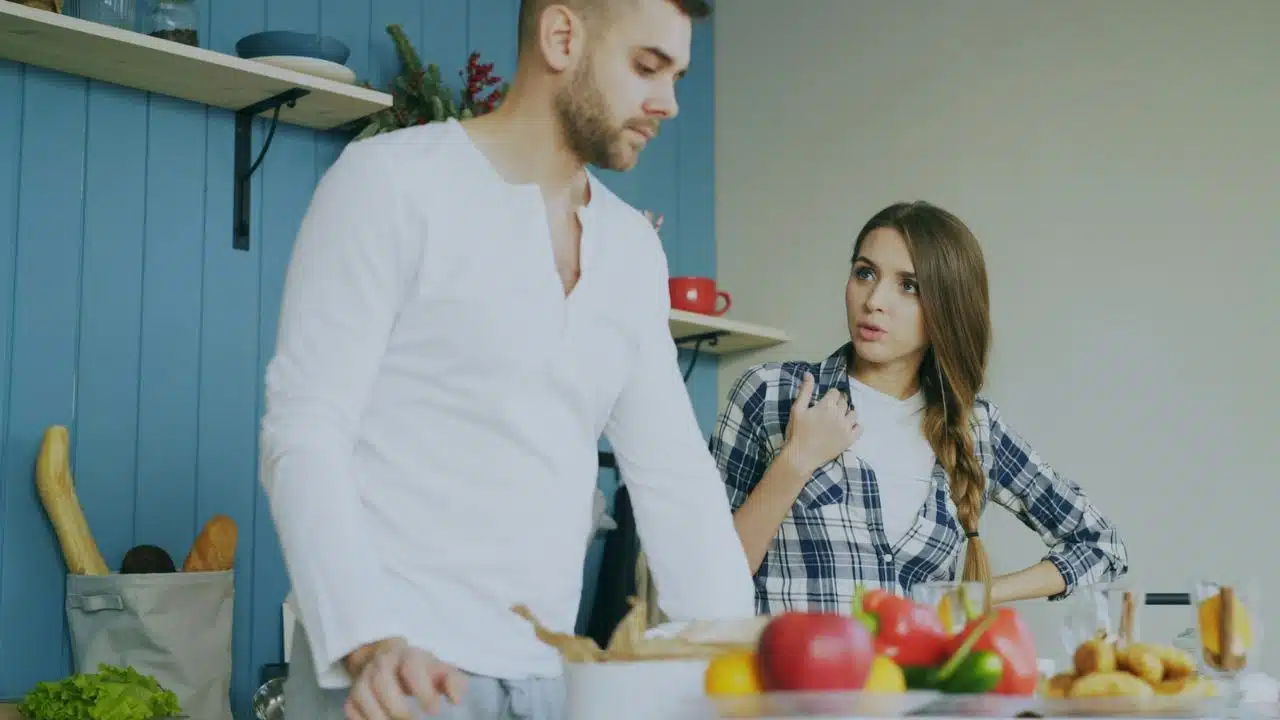 A man stands with his back to a woman who is talking to him in a kitchen.