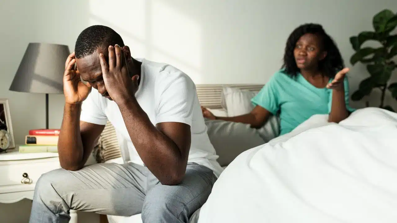 A distressed man sits on a bed holding his head while a woman argues in the background.