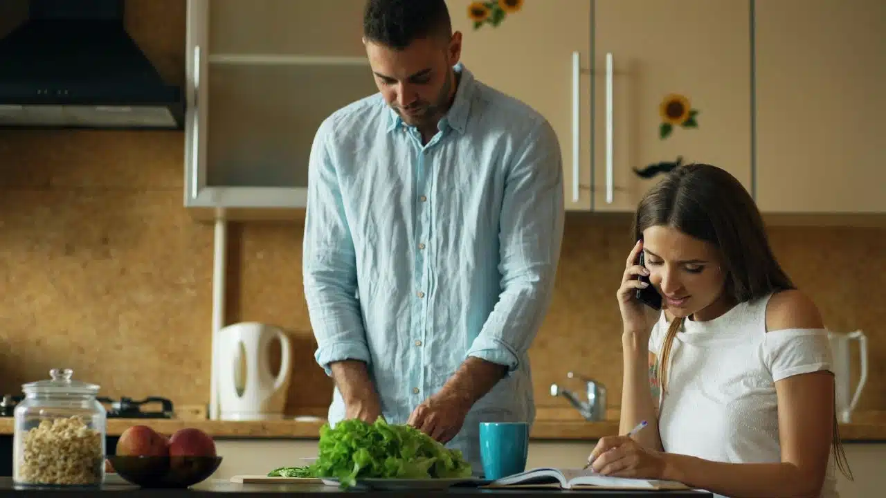 A man chops vegetables while a woman writes and talks on the phone in a kitchen.