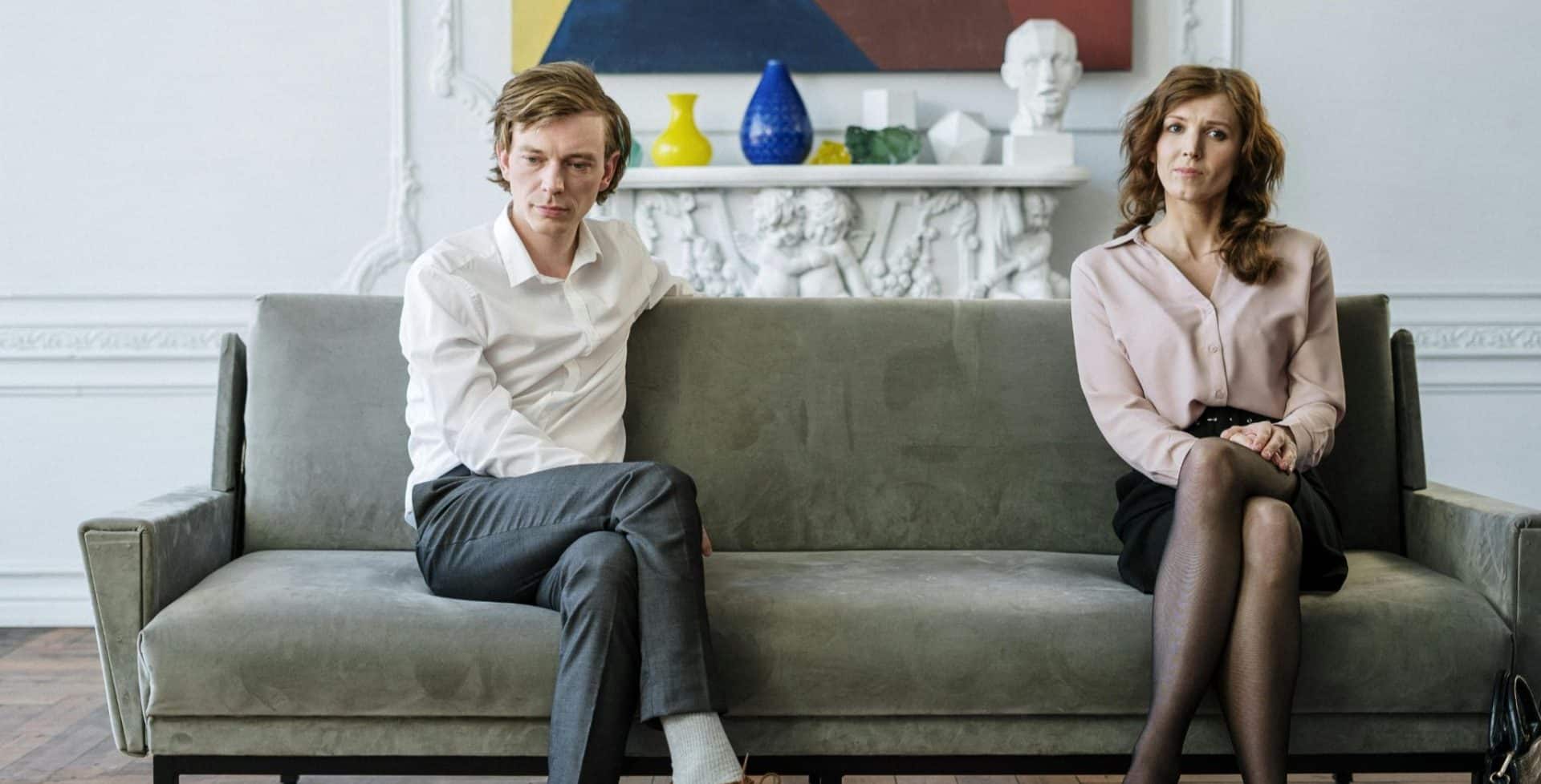 A couple sitting apart on a couch, both looking thoughtful but distant.