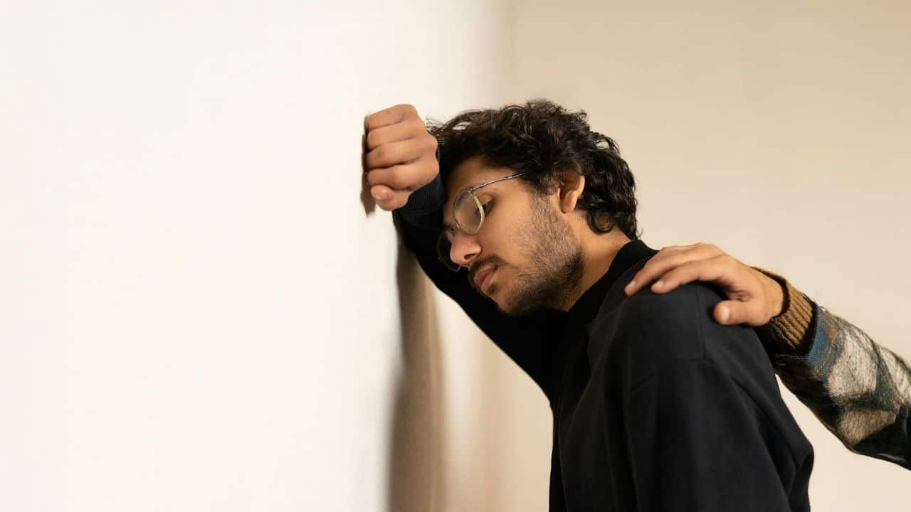 A man leans his head against a white wall while a hand rests on his shoulder.