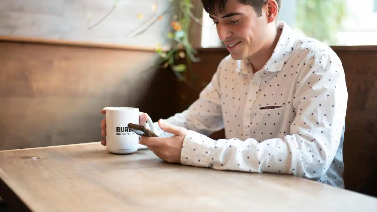 A man sitting at a café table, smiling while looking at his phone and holding a coffee mug.