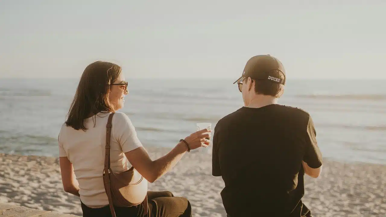 A couple sitting on a beach ledge, sharing a drink while looking out at the ocean.