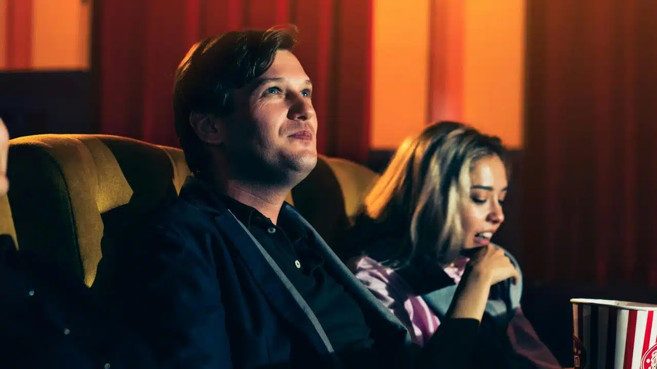 A couple sitting in a movie theater, watching the screen with popcorn nearby.
