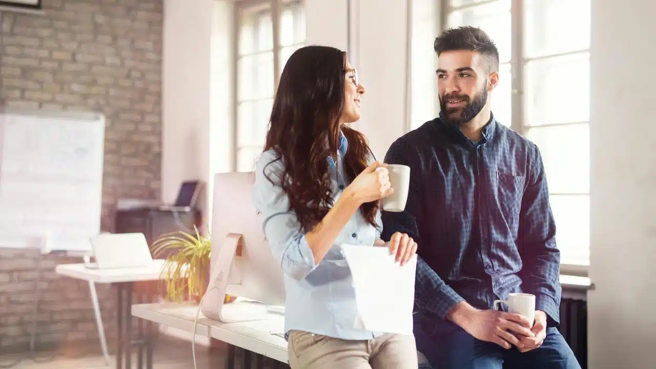 A man and woman chatting in a bright office while holding coffee mugs.