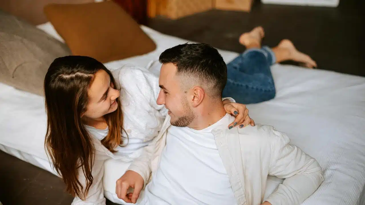 A couple lying on a bed, smiling at each other in a relaxed indoor setting.