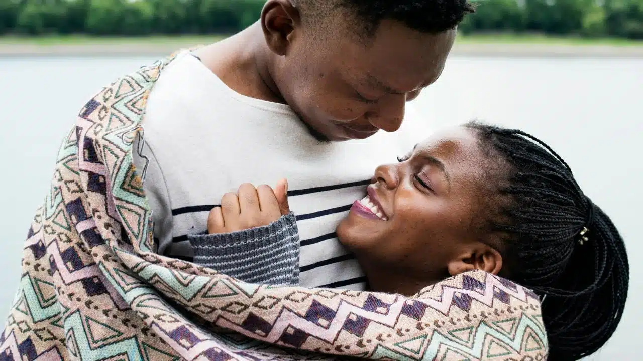 A couple wrapped in a blanket, smiling warmly while embracing outdoors near water.