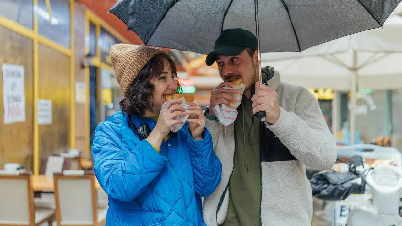 A couple standing under an umbrella in the rain, smiling while eating sandwiches outdoors.