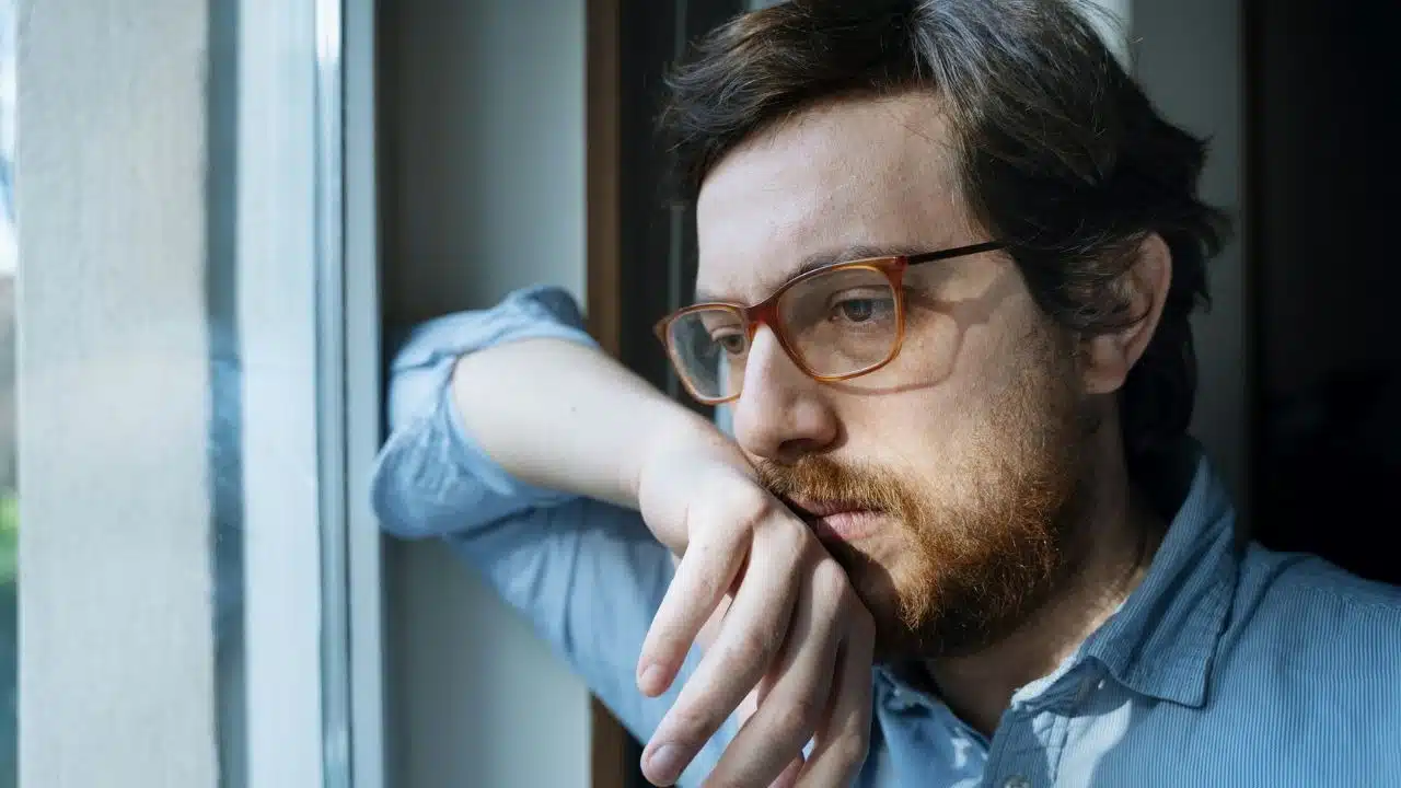 A bearded man wearing glasses leans his arm against a window and looks outside.