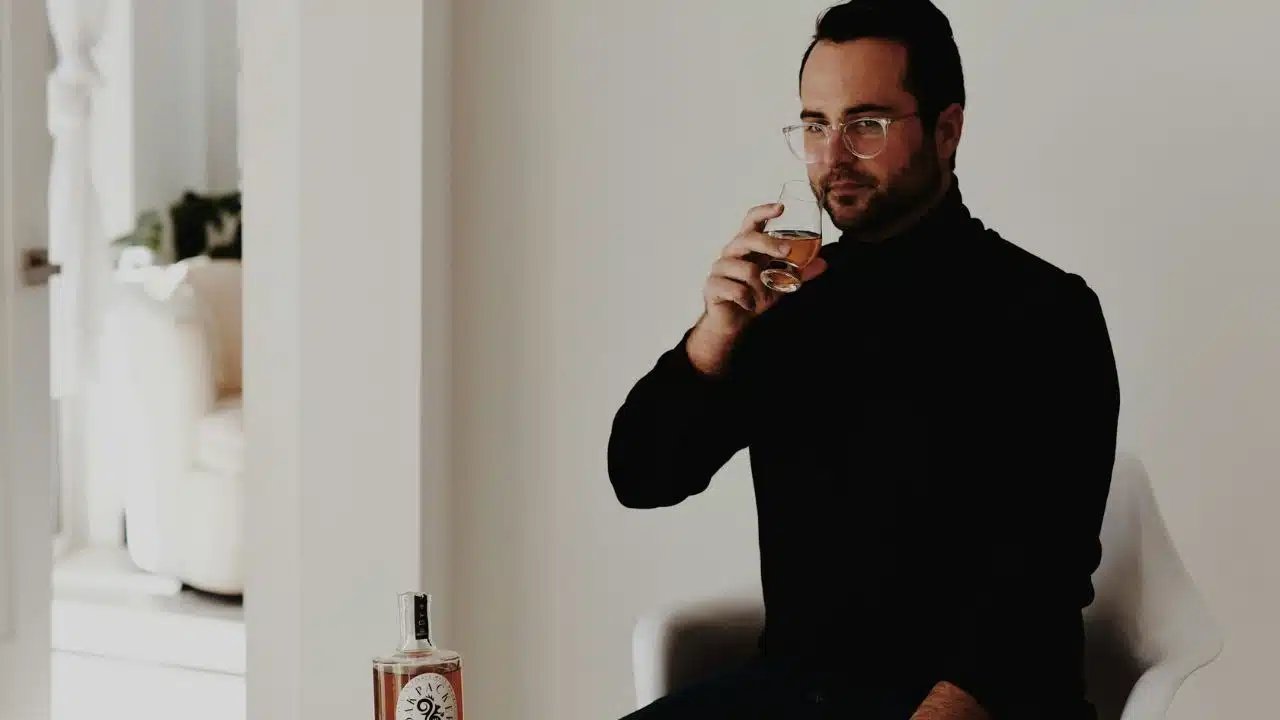 A man with glasses and a black turtleneck holds a glass of amber liquid.