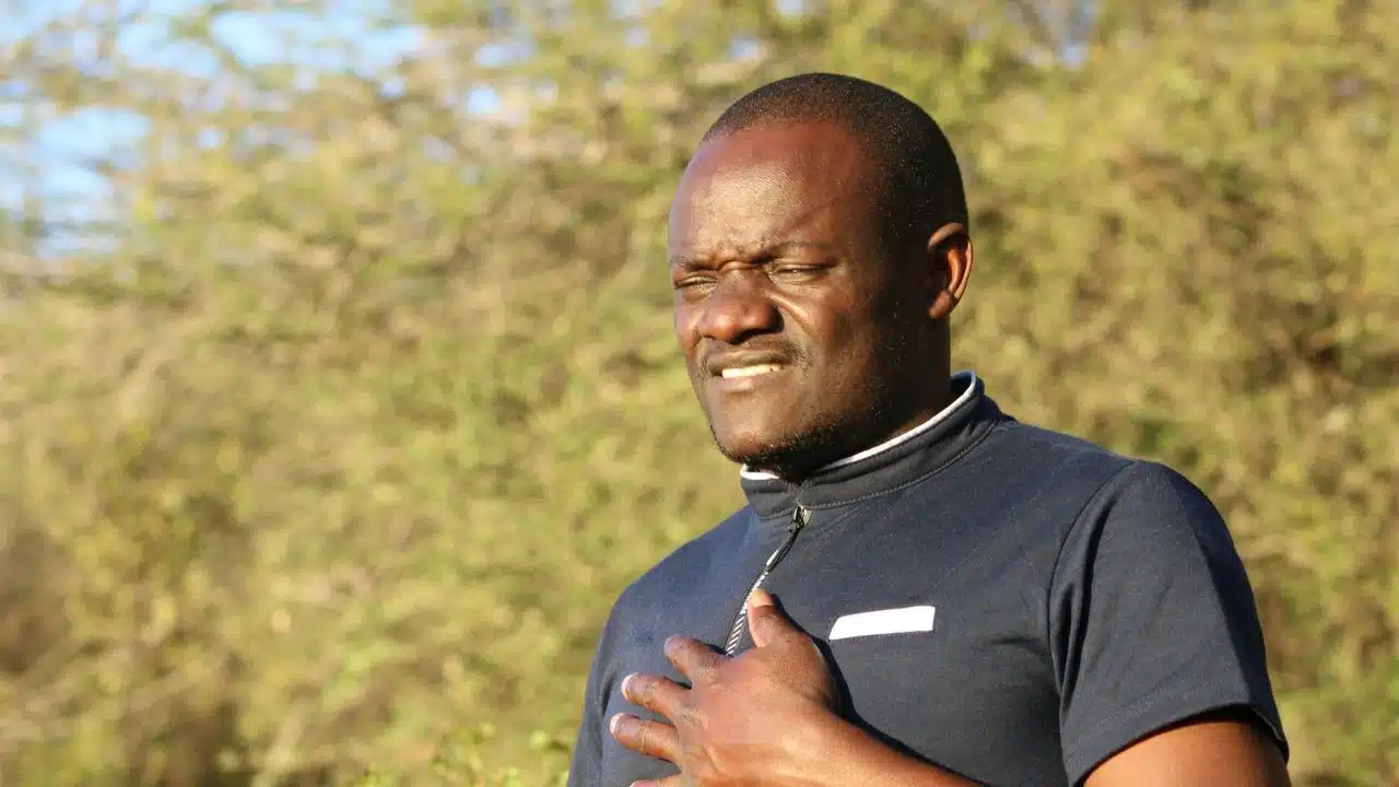 A man in a dark shirt winces while holding his hand against his chest outdoors.