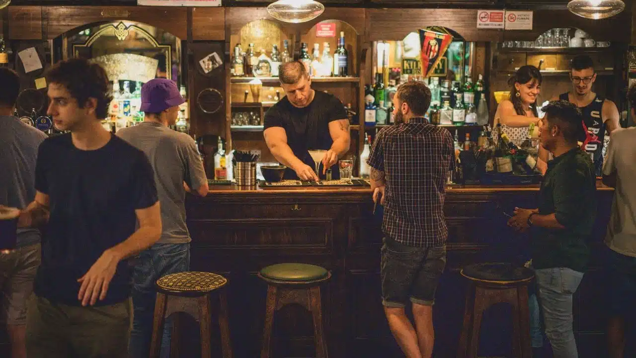 A busy bar scene with a bartender preparing a drink for several patrons standing nearby.
