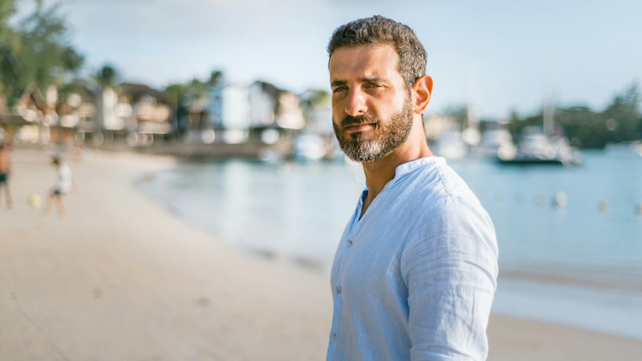 A bearded man looking at the camera while standing on a beach.
