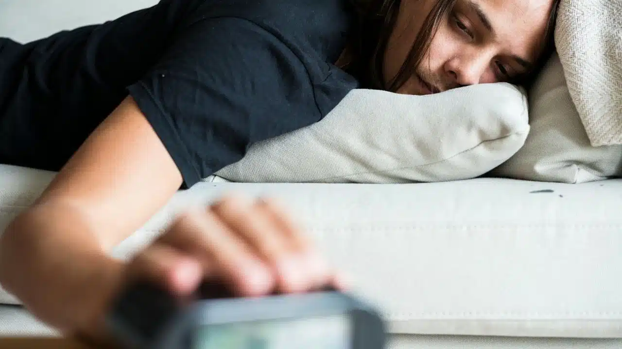 A person lies face down on a pillow while reaching a hand toward an alarm clock.