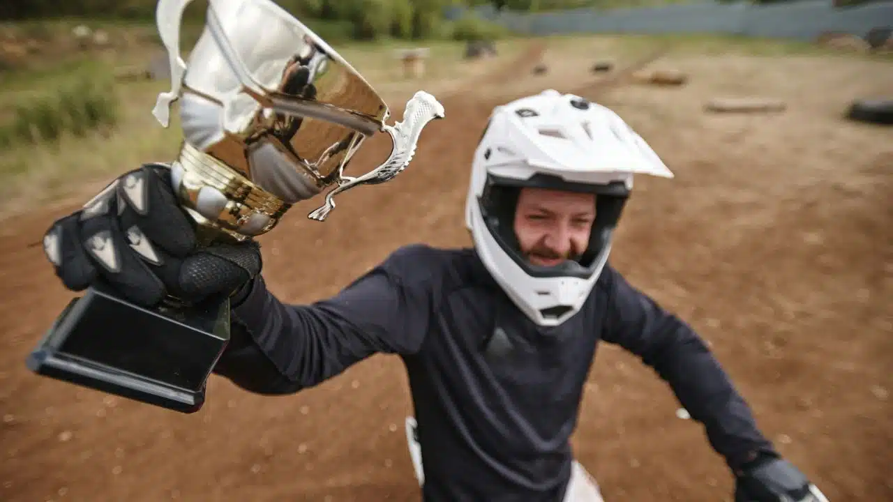 A person in a white motocross helmet and gloves holds up a silver trophy outdoors.