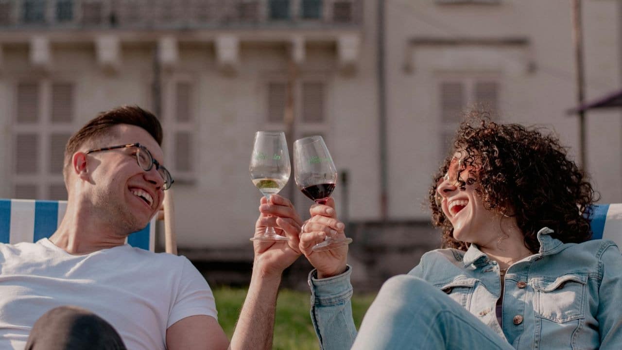 A laughing man and woman clink wine glasses while sitting outdoors in lounge chairs.