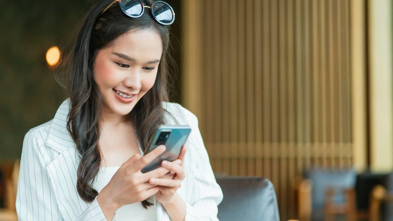 A smiling woman in a white striped blazer looks down at a handheld smartphone.