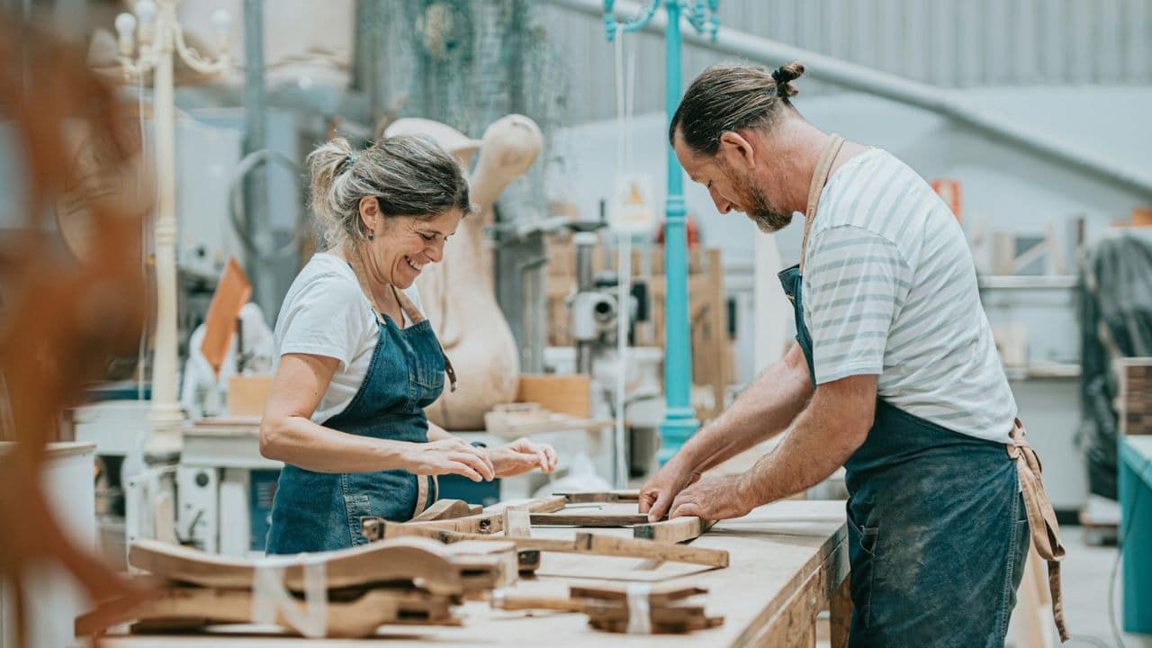 A man and woman wearing denim aprons work together on wooden furniture in a workshop.