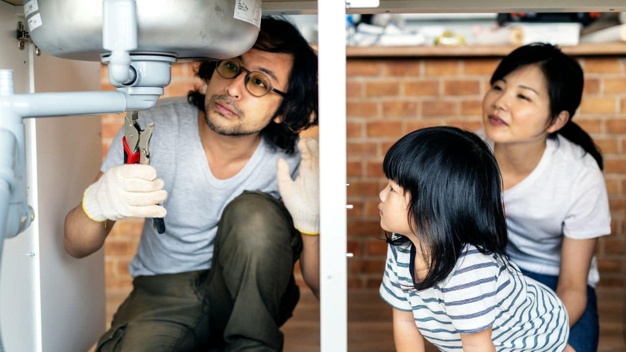 A man uses pliers under a sink while a woman and a young child watch closely.
