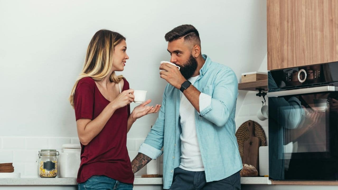 A woman in a red shirt talks to a bearded man drinking from a white cup.