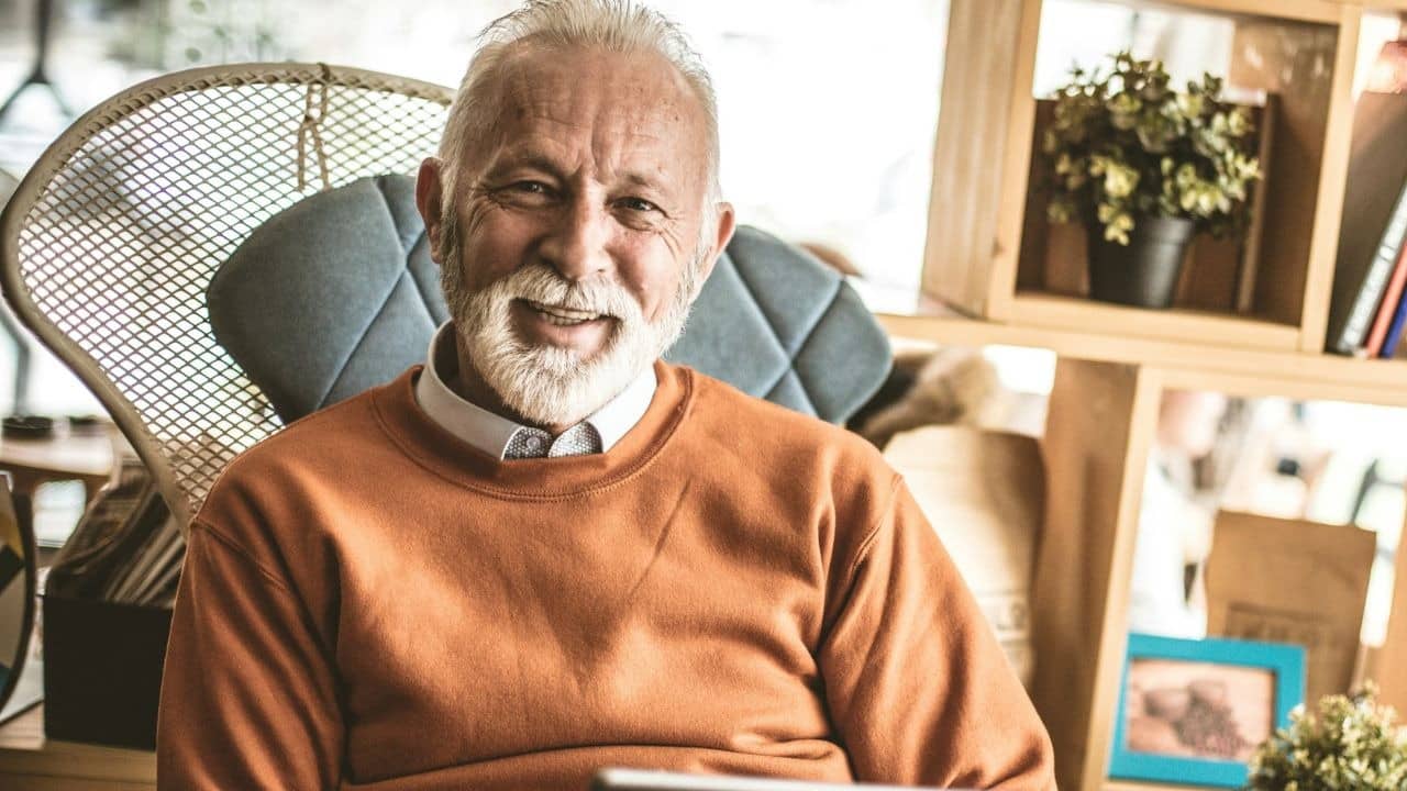 A smiling older man with a white beard sits in a chair wearing an orange sweater.