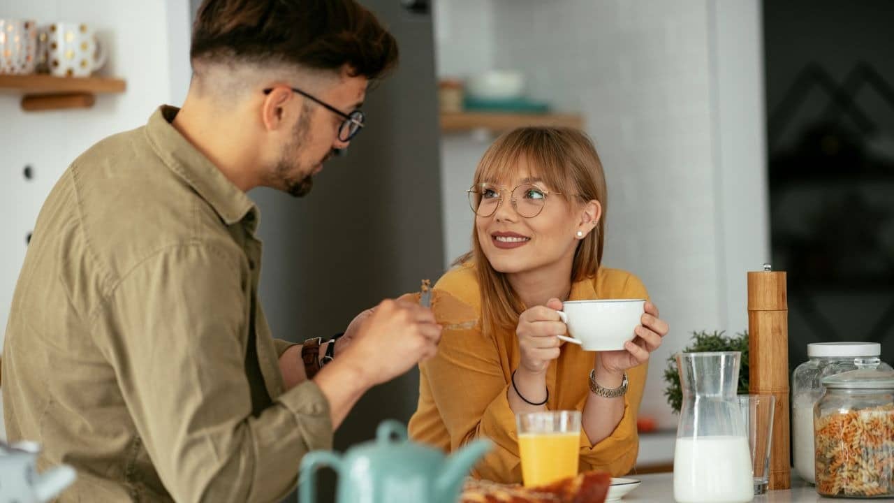 A woman with glasses holds a white mug while looking at a man eating breakfast.