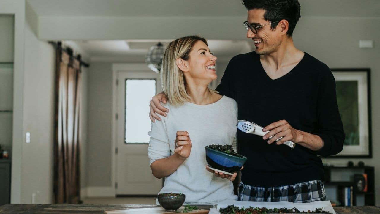 A man with his arm around a woman holds kitchen tongs while they both smile.