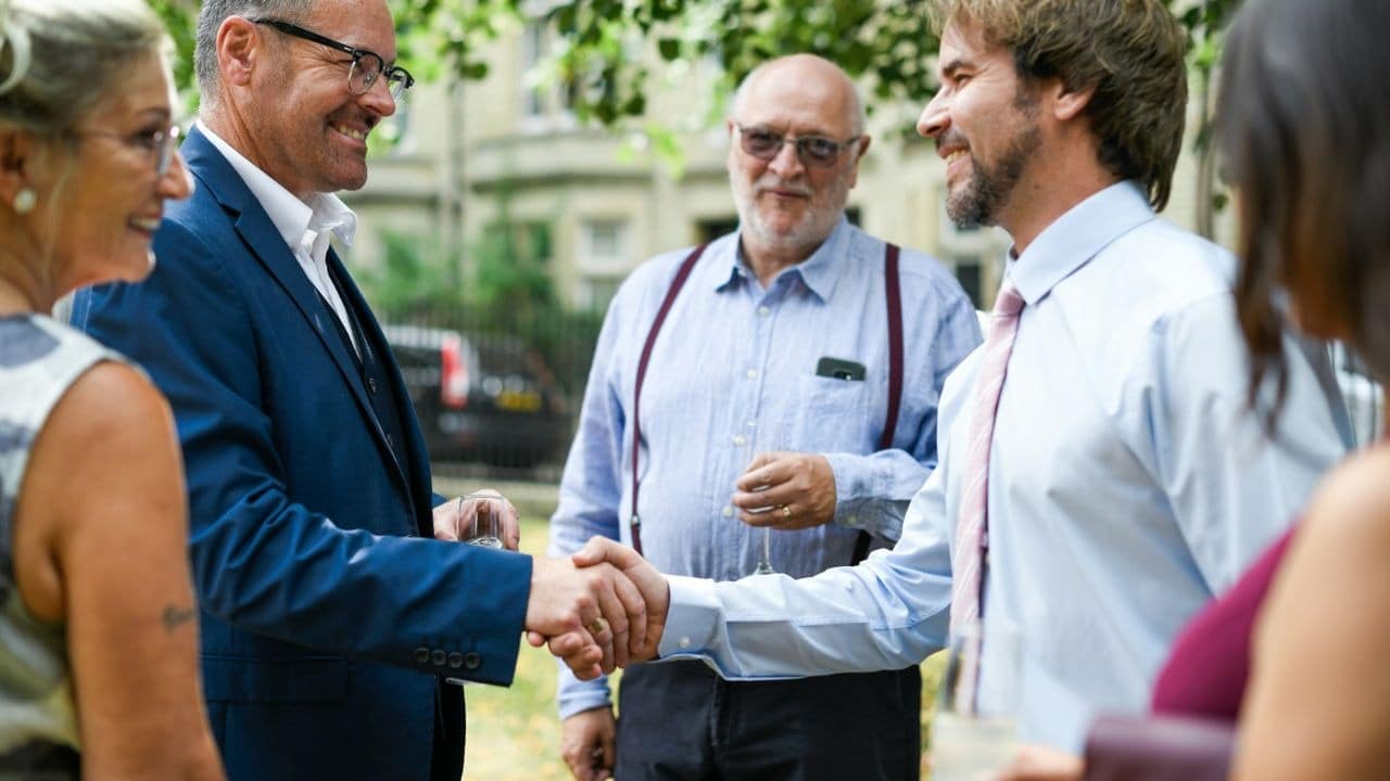 Two men in formal attire shake hands while other people stand nearby in an outdoor setting.